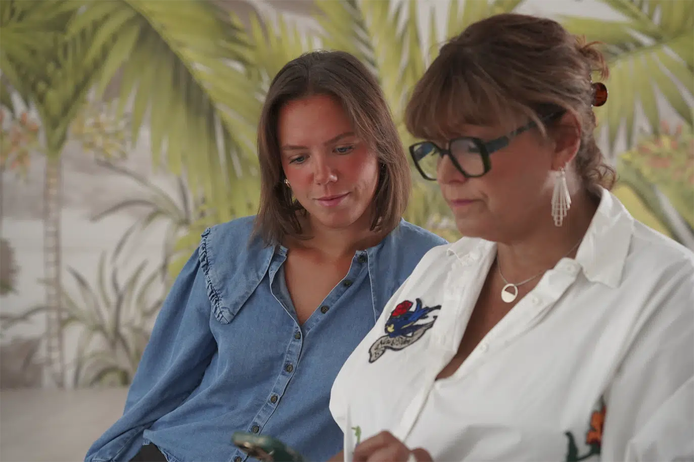 eux femmes de générations différentes regardent attentivement l'écran d'un smartphone ensemble, devant un fond aux motifs de feuilles tropicales.